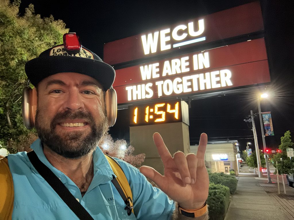 I_Am_Getting_Married_v1.zip 14 Selfie of Tukayote at night under a WECU sign that reads We Are In This Together