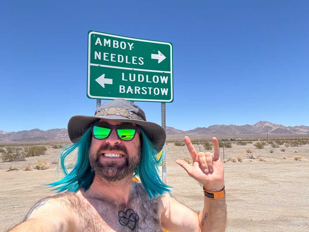 I_Am_Getting_Married_v1.zip 16 Shirtless selfie of Tukayote with teal hair under an Amboy, Needles, Ludlow, and Barstow road sign in the desert