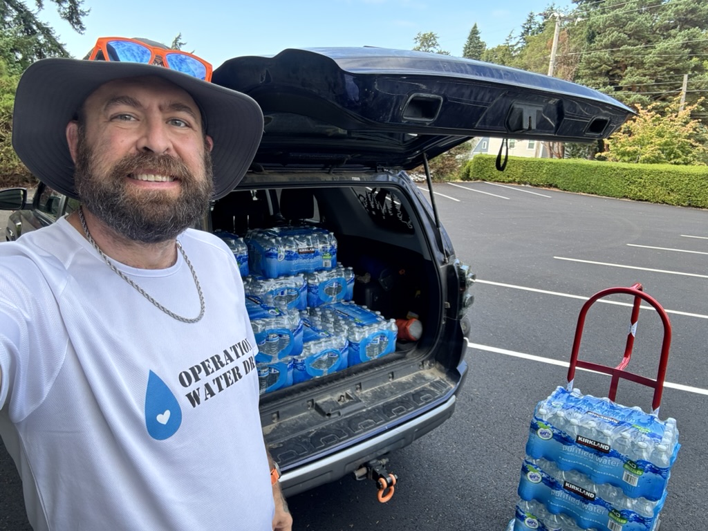 Selfie of Tukayote in an Operation Water shirt beside an SUV loaded with bottled water