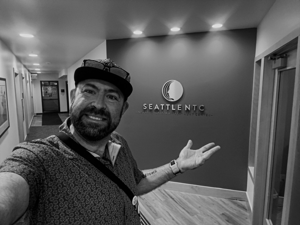 Black and white selfie of Tukayote gesturing toward a Seattle ENT clinic sign