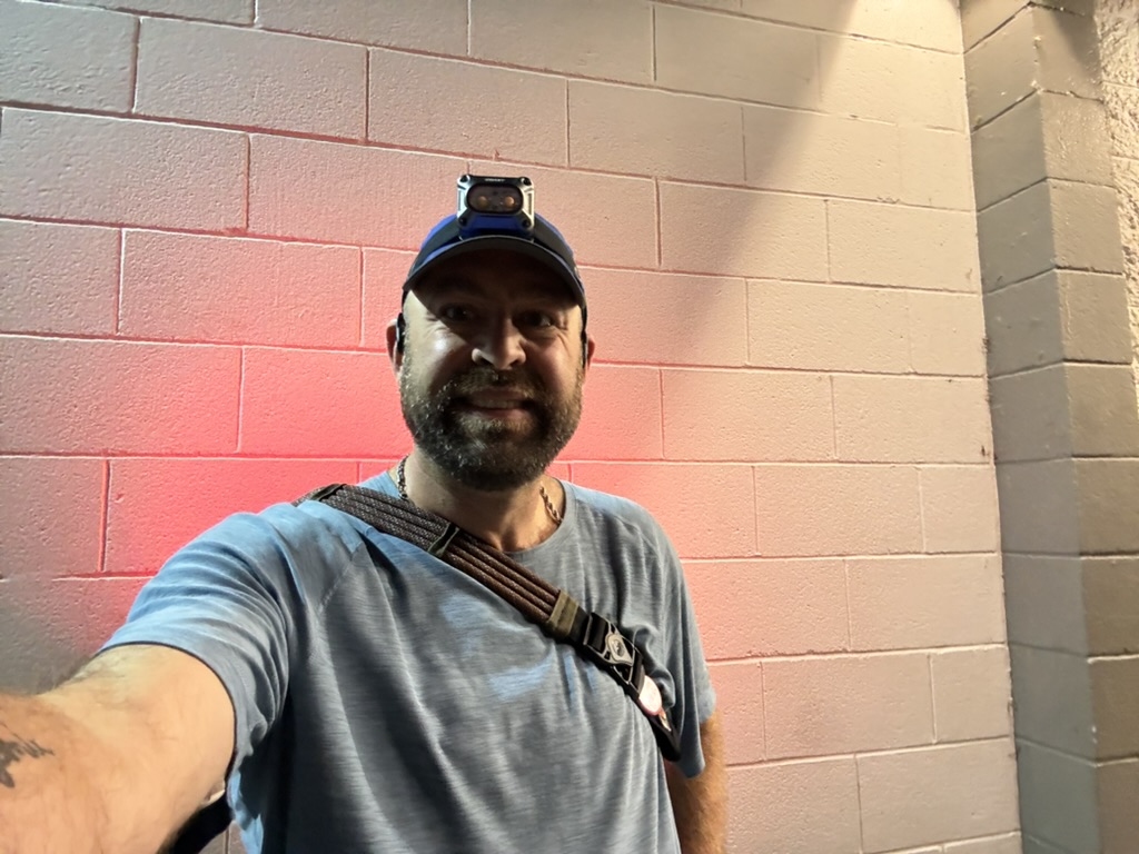 Selfie of Tukayote in a cap against a cinder block wall lit pink