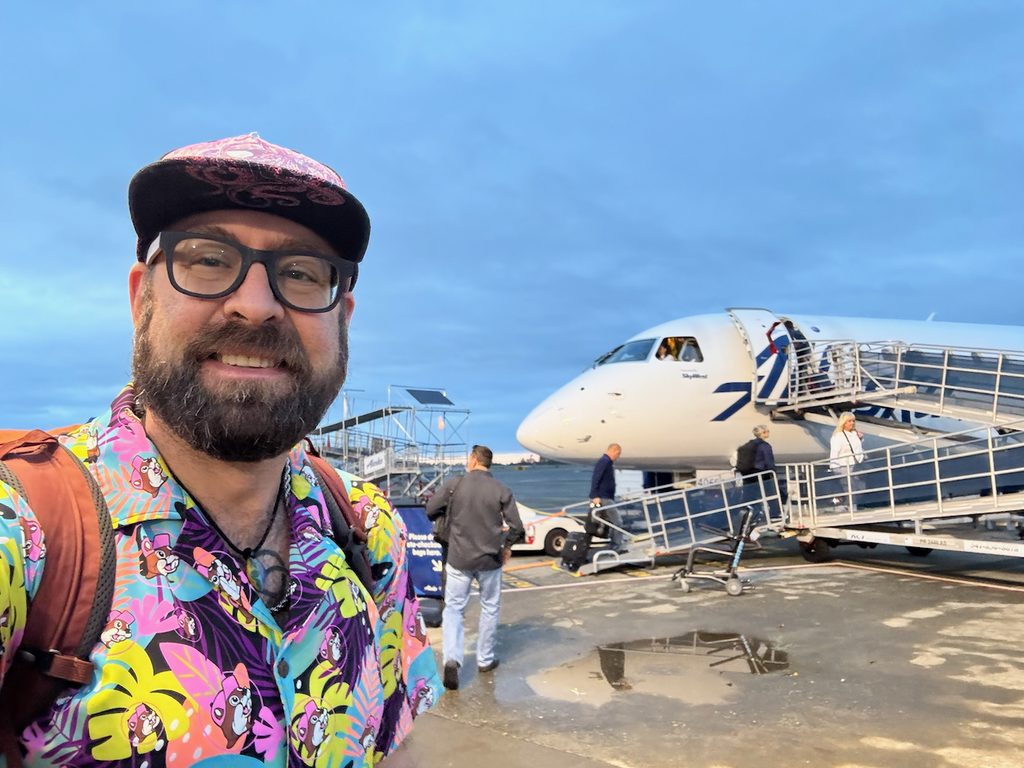 Selfie of Tukayote in a bright tropical shirt in front of a small passenger plane on the tarmac