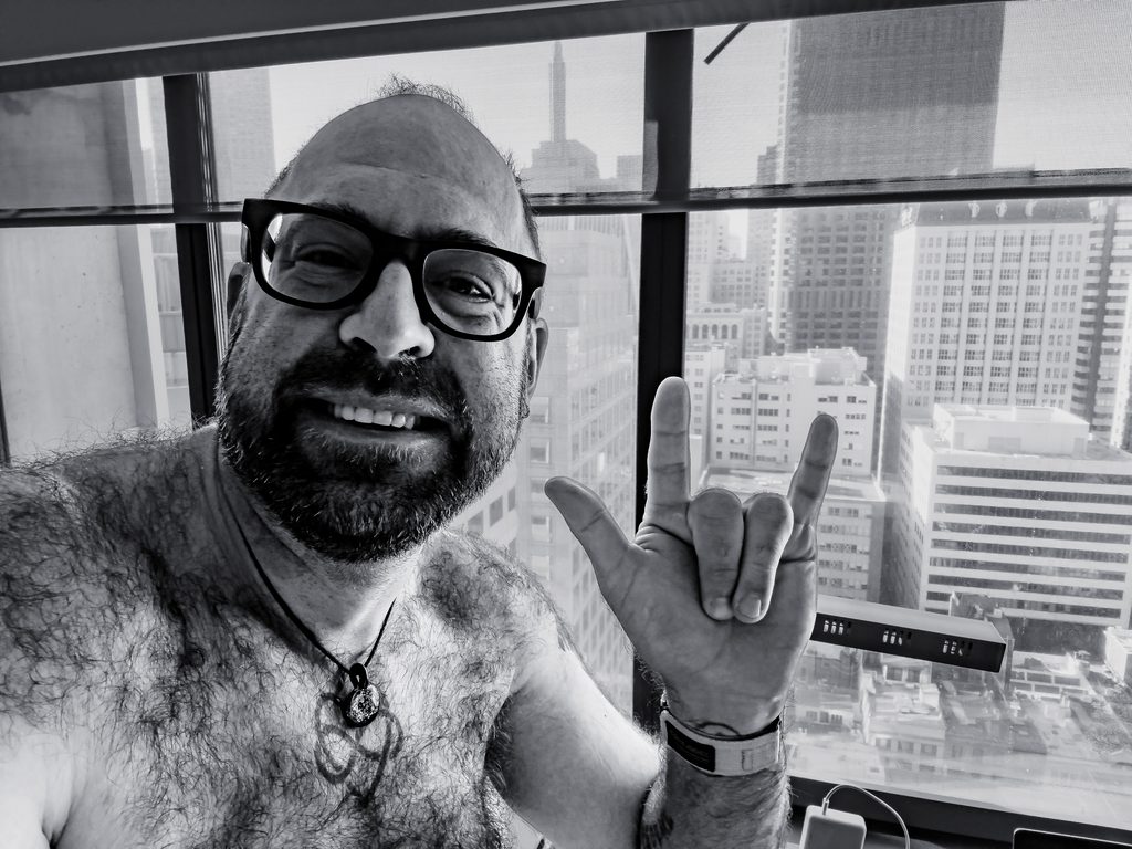 Black and white selfie of Tukayote turning 43, flashing the ASL I love you sign by a high-rise window