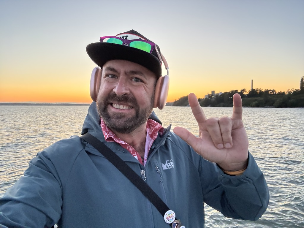 Tukayote smiles in a sunset selfie by the water in a gray-blue jacket, pink shirt, black cap, and pink headphones, making the ASL I love you sign