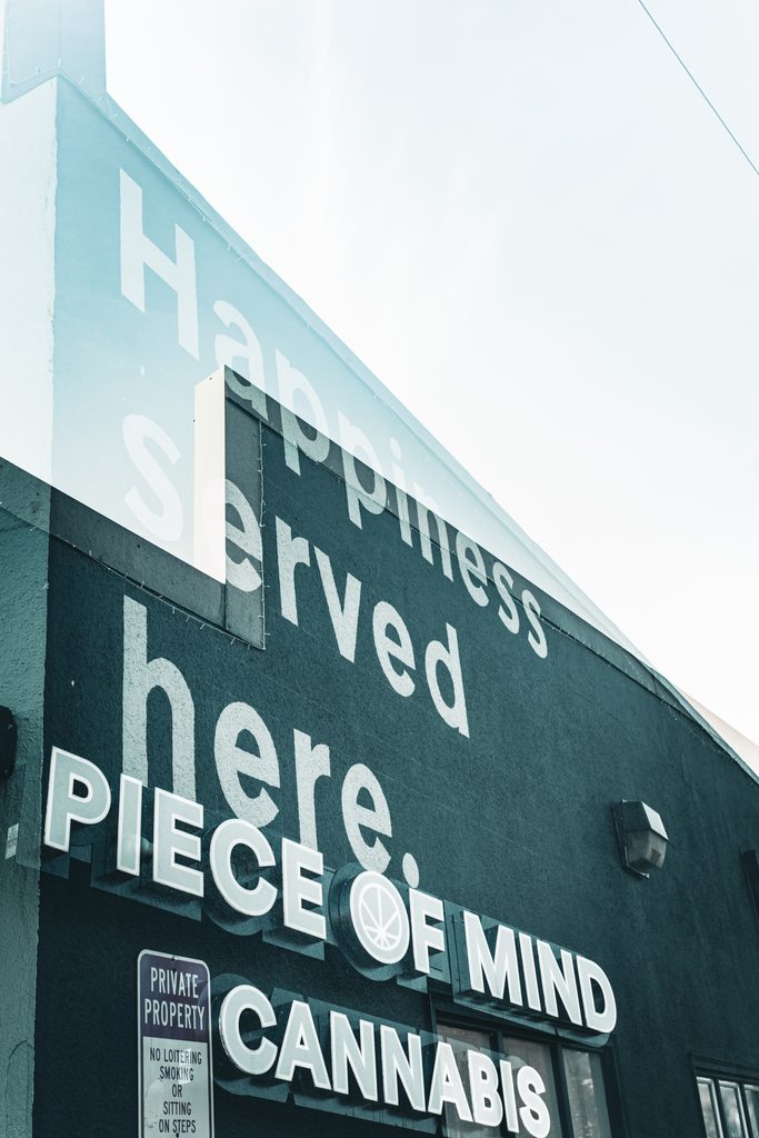 Black building with a large white sign reading Happiness served here, Piece of Mind Cannabis, photographed looking up from the street
