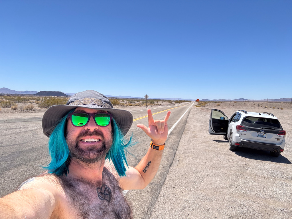 Tukayote takes a bare-chested selfie on an empty desert highway in a wide-brim hat and green mirrored sunglasses, making the ASL I love you sign, with a white SUV and open door behind him