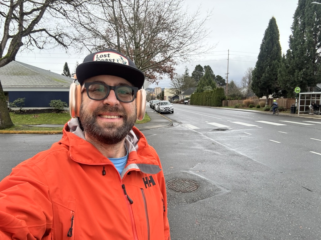 Tukayote takes a selfie on a wet neighborhood street in an orange rain jacket, Lost Coast cap, glasses, and pink headphones