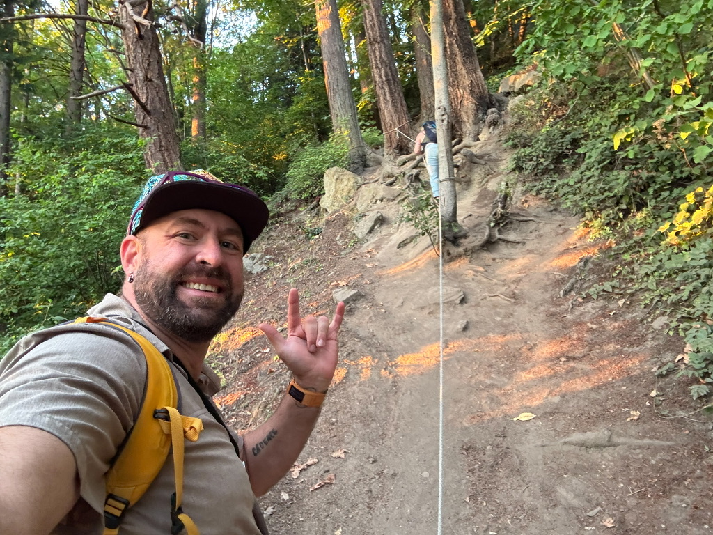 Tukayote smiles on a steep dirt trail through the forest, wearing a yellow backpack and making the ASL I love you sign, with a white rope running down the slope beside him