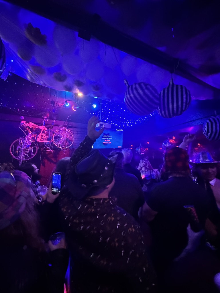 A crowded New Year's Eve party in a dim venue lit blue and purple, with black and white striped lanterns hanging from the ceiling and guests in dark glittery outfits and hats