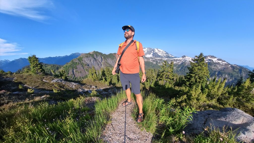 Tukayote stands on a narrow rocky mountain trail in sandals and an orange shirt with a camera across his chest, with snow-capped peaks and evergreens behind him under a clear blue sky