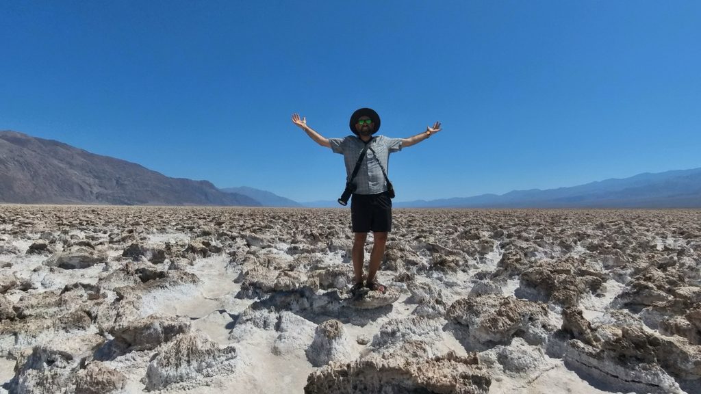 Tukayote stands with arms spread wide on the white salt flats of Devil's Golf Course in Death Valley under a clear blue sky, with brown mountains in the distance