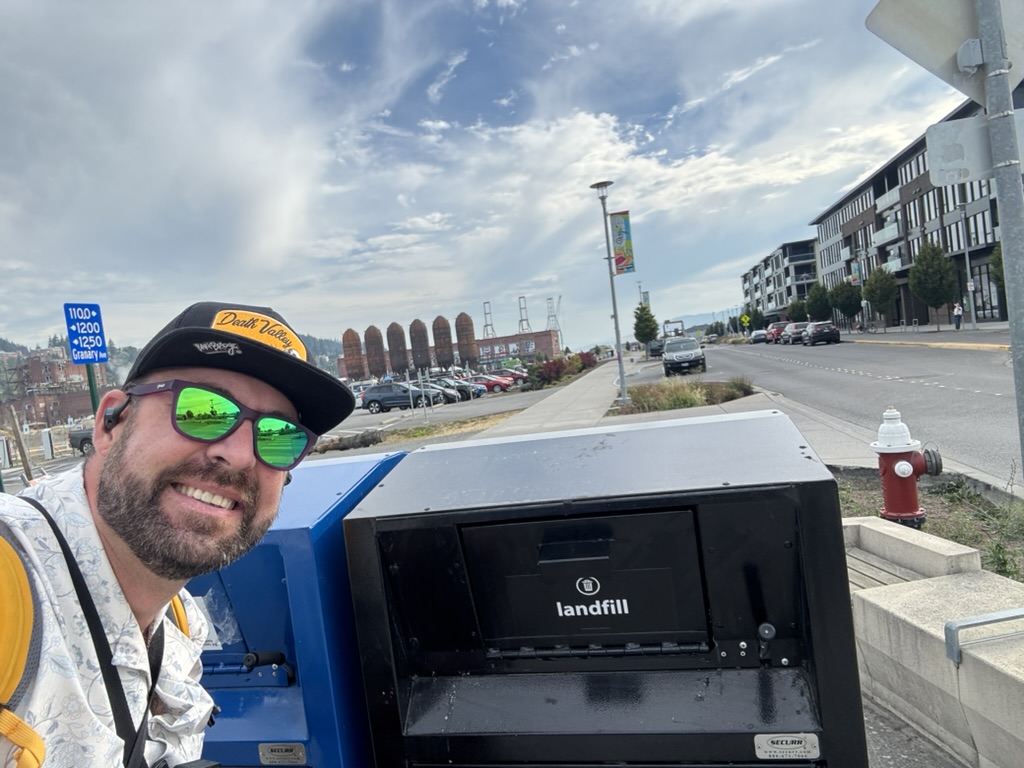 Tukayote takes a smiling selfie beside a black landfill trash bin on the Waterfront near Granary Ave, wearing a Death Valley cap, green mirrored sunglasses, and a patterned shirt
