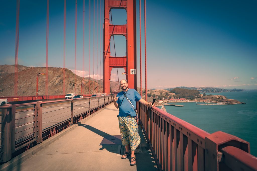 Tukayote smiles on the Golden Gate Bridge walkway in a blue t-shirt and patterned shorts, making the ASL I love you sign, with the red tower and the bay behind him