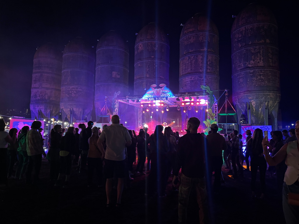 A crowd watches Reposado perform on a lit stage at night in front of tall concrete grain silos at the Trackside Mural Festival