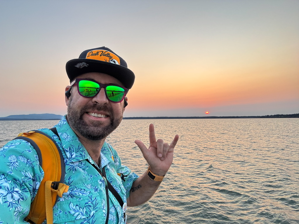 Tukayote takes a sunset selfie over calm water, smiling in a Death Valley cap, green mirrored sunglasses, and a turquoise Hawaiian shirt and making the ASL I love you sign