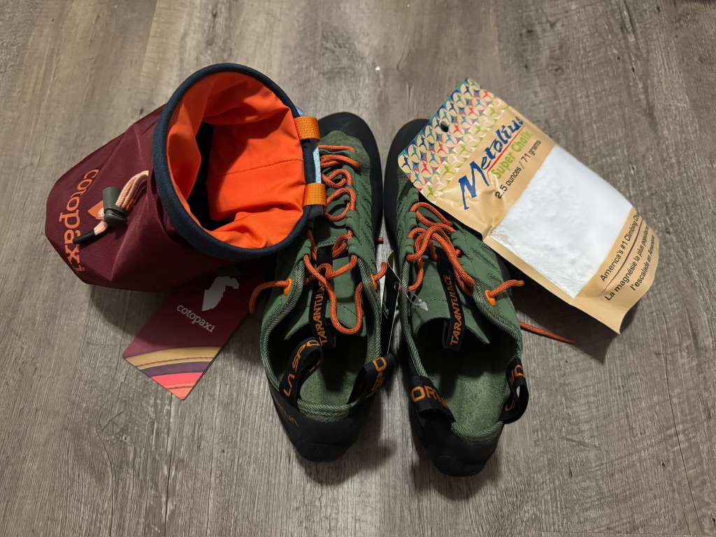 Climbing gear on a wood floor: a maroon chalk bag, green and orange climbing shoes, and a bag of climbing chalk