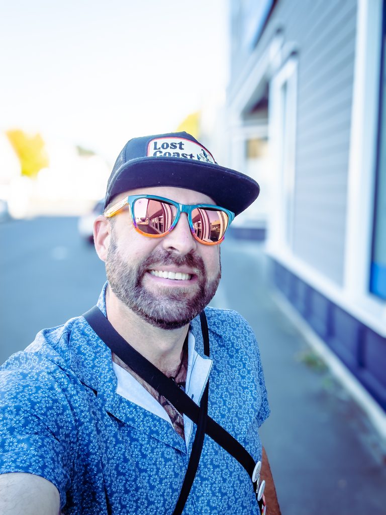 Close-up selfie of Tukayote, chemically sober and smiling in a Lost Coast trucker cap, rainbow mirrored sunglasses, and a blue floral shirt on a sunny street