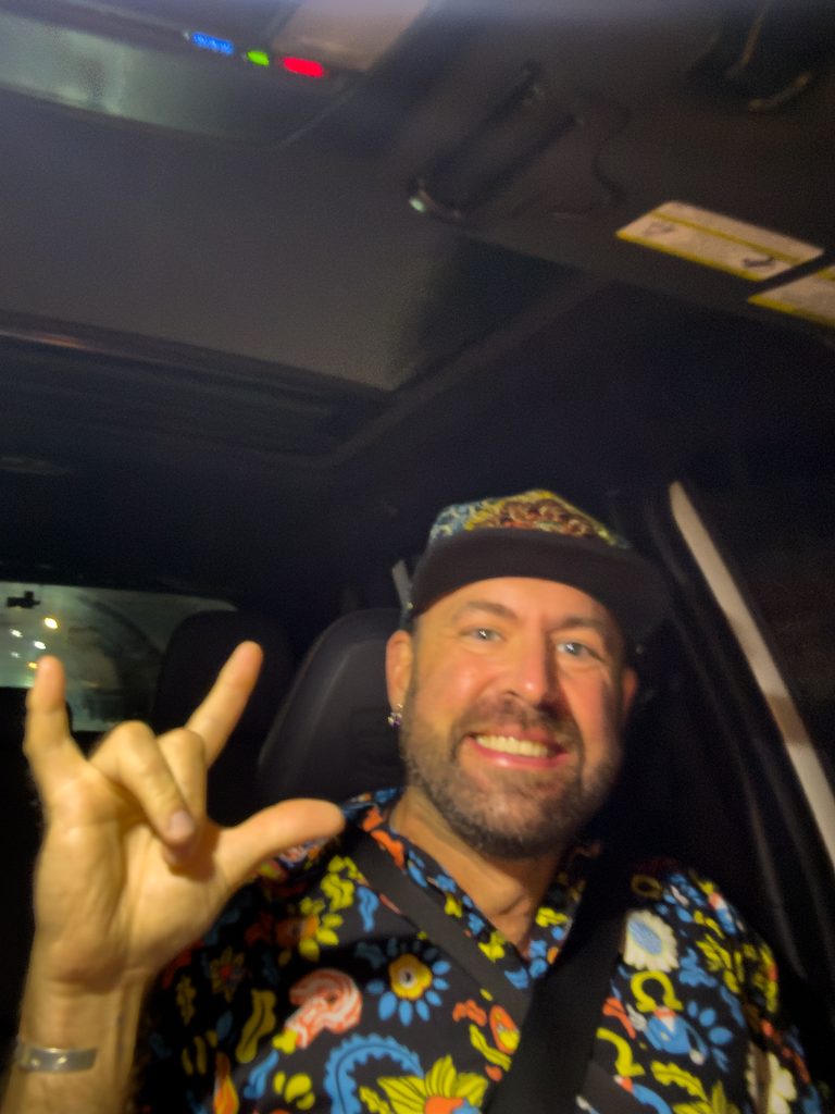 Tukayote smiling in a car at night in a colorful floral shirt and black cap, making the ASL I love you sign on his way to a rave