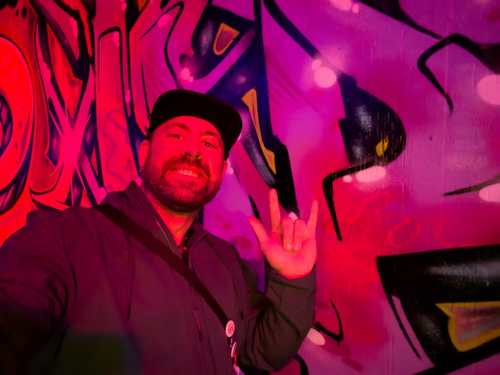 Tukayote takes a selfie in front of a glowing pink and red graffiti mural, wearing a black cap and dark jacket and making the ASL I love you sign